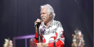 Joe Bonsall with white hair, wearing a festive shirt with musical notes and Santa Claus faces, sings into a microphone on stage.