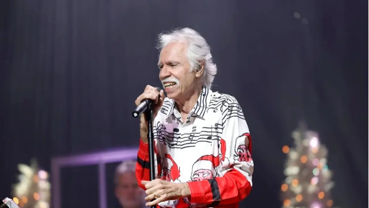 Joe Bonsall with white hair, wearing a festive shirt with musical notes and Santa Claus faces, sings into a microphone on stage.