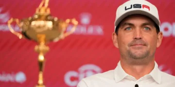 A man in a white shirt and cap, identified as U.S. Ryder Cup Captain Keegan Bradley, stands next to a golden trophy against a red backdrop, possibly celebrating the 2025 event.
