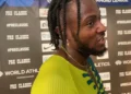Kishane Thompson with braided hair and a beard, stands in front of a backdrop featuring various sponsors' logos. Wearing a yellow and blue athletic top, he seems to be speaking or reacting, likely after competing in the 100m sprint at the Jamaican Olympic Trials.
