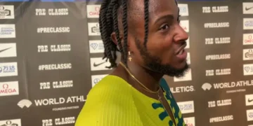 Kishane Thompson with braided hair and a beard, stands in front of a backdrop featuring various sponsors' logos. Wearing a yellow and blue athletic top, he seems to be speaking or reacting, likely after competing in the 100m sprint at the Jamaican Olympic Trials.