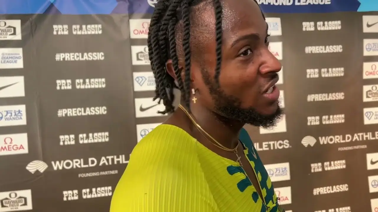 Kishane Thompson with braided hair and a beard, stands in front of a backdrop featuring various sponsors' logos. Wearing a yellow and blue athletic top, he seems to be speaking or reacting, likely after competing in the 100m sprint at the Jamaican Olympic Trials.