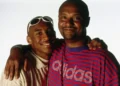 Two individuals standing close together, smiling at the camera with arms around each other's shoulders. One wears sunglasses on his head and the other a striped Adidas shirt, reminiscent of a photograph of Kobe Bryant and his father Joe Jellybean Bryant.