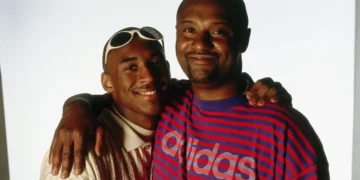 Two individuals standing close together, smiling at the camera with arms around each other's shoulders. One wears sunglasses on his head and the other a striped Adidas shirt, reminiscent of a photograph of Kobe Bryant and his father Joe Jellybean Bryant.