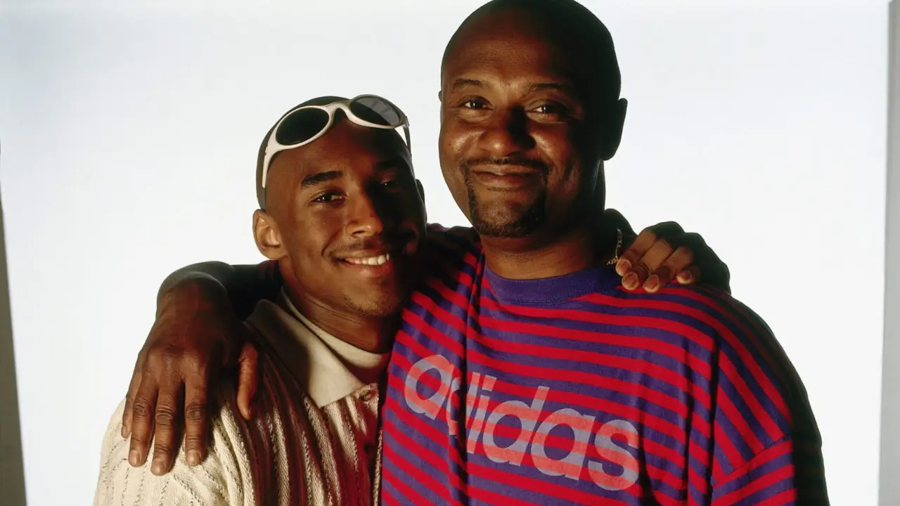 Two individuals standing close together, smiling at the camera with arms around each other's shoulders. One wears sunglasses on his head and the other a striped Adidas shirt, reminiscent of a photograph of Kobe Bryant and his father Joe Jellybean Bryant.