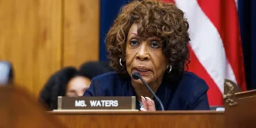 Maxine Waters with curly hair sits at a desk with a nameplate reading "Ms. Waters," speaking into a microphone, with a U.S. flag in the background. It appears to be an interview for CNN, featuring Congresswoman Maxine Waters.