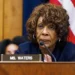 Maxine Waters with curly hair sits at a desk with a nameplate reading "Ms. Waters," speaking into a microphone, with a U.S. flag in the background. It appears to be an interview for CNN, featuring Congresswoman Maxine Waters.