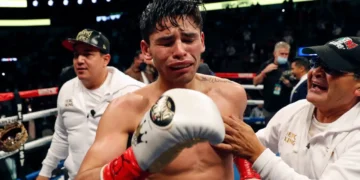 Ryan Garcia is visibly emotional in the ring, flanked by two team members, with one placing a hand on the boxer's chest. The scene is set in a well-lit arena filled with spectators.