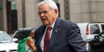 Sen.-Bob-Menendez with white hair and glasses, dressed in a dark suit and red tie, is walking outside in a city setting with other people and vehicles in the background. This scene unfolds as he arrives for Sen. Bob Menendez's federal corruption trial.