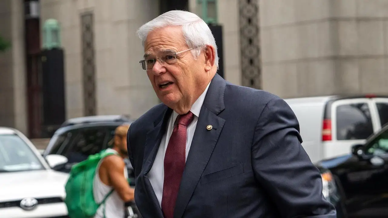 Sen.-Bob-Menendez with white hair and glasses, dressed in a dark suit and red tie, is walking outside in a city setting with other people and vehicles in the background. This scene unfolds as he arrives for Sen. Bob Menendez's federal corruption trial.