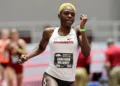 Shafiqua Maloney with short blonde hair competes in an 800m running event, wearing a white Arkansas uniform and a bib labeled "Shafiqua Maloney." Others in similar attire are blurred in the background, hinting at the fierce competition leading up to the Paris 2024 Olympics.