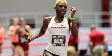 Shafiqua Maloney with short blonde hair competes in an 800m running event, wearing a white Arkansas uniform and a bib labeled "Shafiqua Maloney." Others in similar attire are blurred in the background, hinting at the fierce competition leading up to the Paris 2024 Olympics.