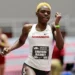 Shafiqua Maloney with short blonde hair competes in an 800m running event, wearing a white Arkansas uniform and a bib labeled "Shafiqua Maloney." Others in similar attire are blurred in the background, hinting at the fierce competition leading up to the Paris 2024 Olympics.