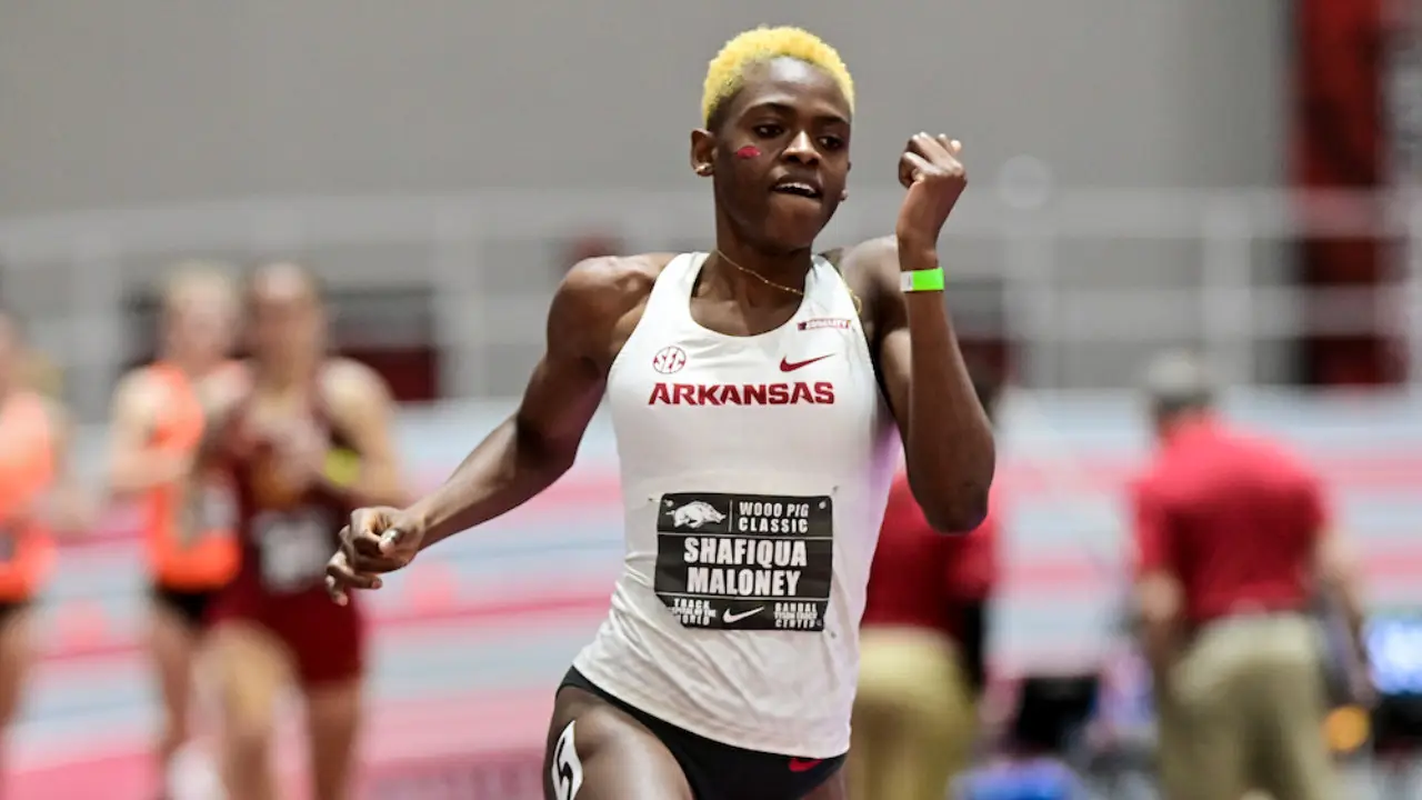 Shafiqua Maloney with short blonde hair competes in an 800m running event, wearing a white Arkansas uniform and a bib labeled "Shafiqua Maloney." Others in similar attire are blurred in the background, hinting at the fierce competition leading up to the Paris 2024 Olympics.