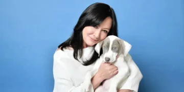 Shannen Doherty, Star of '90210' and 'Charmed,' Dies at 53 After Battle with Cancer with long black hair, wearing a white outfit, holds a white and gray puppy against a blue background.