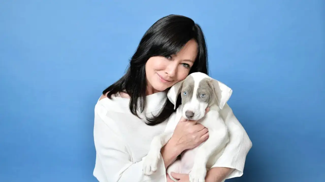 Shannen Doherty, Star of '90210' and 'Charmed,' Dies at 53 After Battle with Cancer with long black hair, wearing a white outfit, holds a white and gray puppy against a blue background.