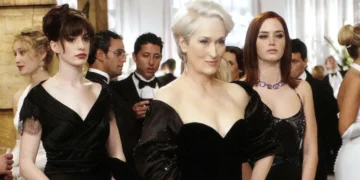 Three women in elegant black dresses stand in a formal event setting. Several other formally dressed individuals are in the background, including a man in a tuxedo. Rumors swirl about Emily Blunt and Anne Hathaway possibly discussing "The Devil Wears Prada" sequel at the event.