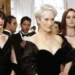 Three women in elegant black dresses stand in a formal event setting. Several other formally dressed individuals are in the background, including a man in a tuxedo. Rumors swirl about Emily Blunt and Anne Hathaway possibly discussing "The Devil Wears Prada" sequel at the event.