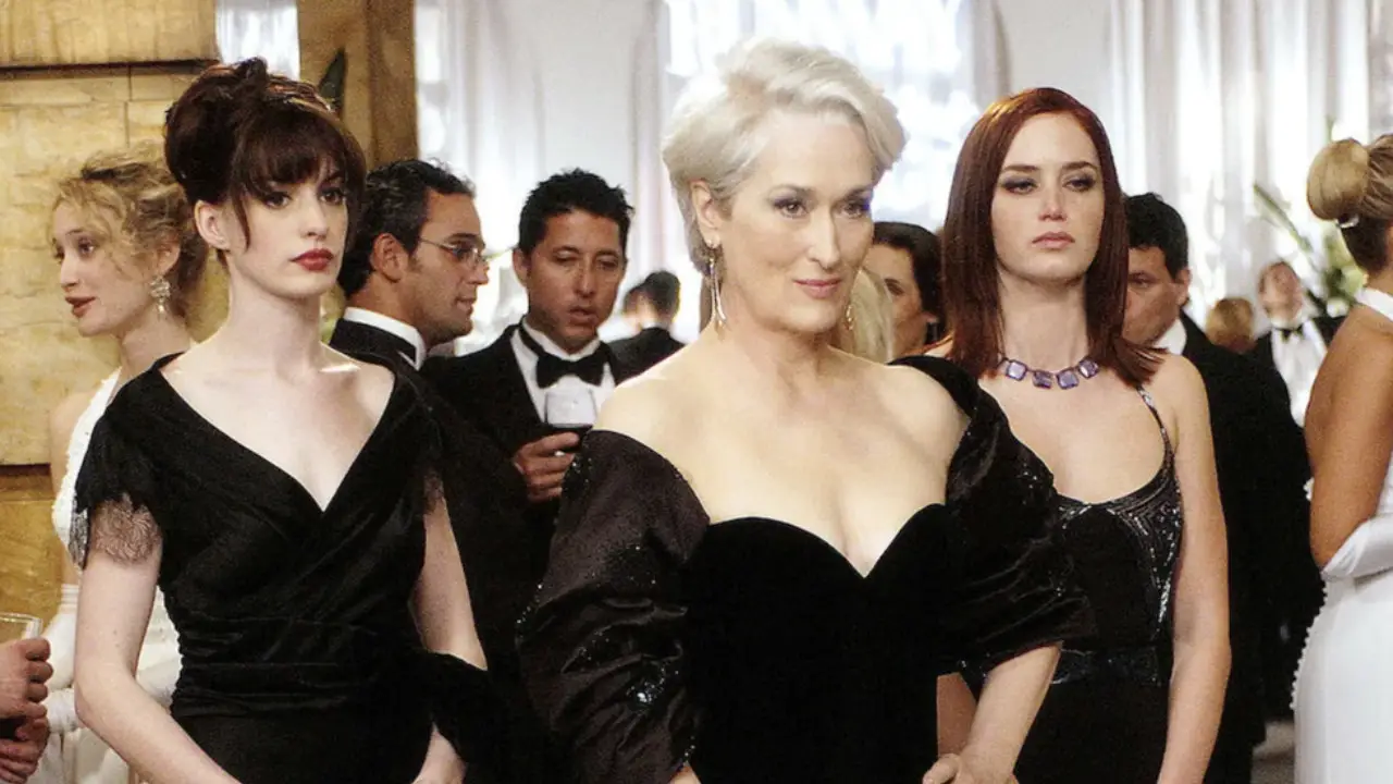 Three women in elegant black dresses stand in a formal event setting. Several other formally dressed individuals are in the background, including a man in a tuxedo. Rumors swirl about Emily Blunt and Anne Hathaway possibly discussing "The Devil Wears Prada" sequel at the event.