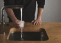 A person is holding a glass under a running faucet, letting water overflow into a modern wooden sink, mindful of Ofwat regulations.