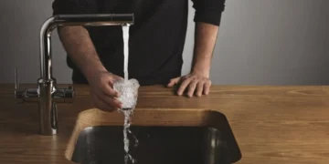 A person is holding a glass under a running faucet, letting water overflow into a modern wooden sink, mindful of Ofwat regulations.