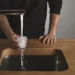 A person is holding a glass under a running faucet, letting water overflow into a modern wooden sink, mindful of Ofwat regulations.