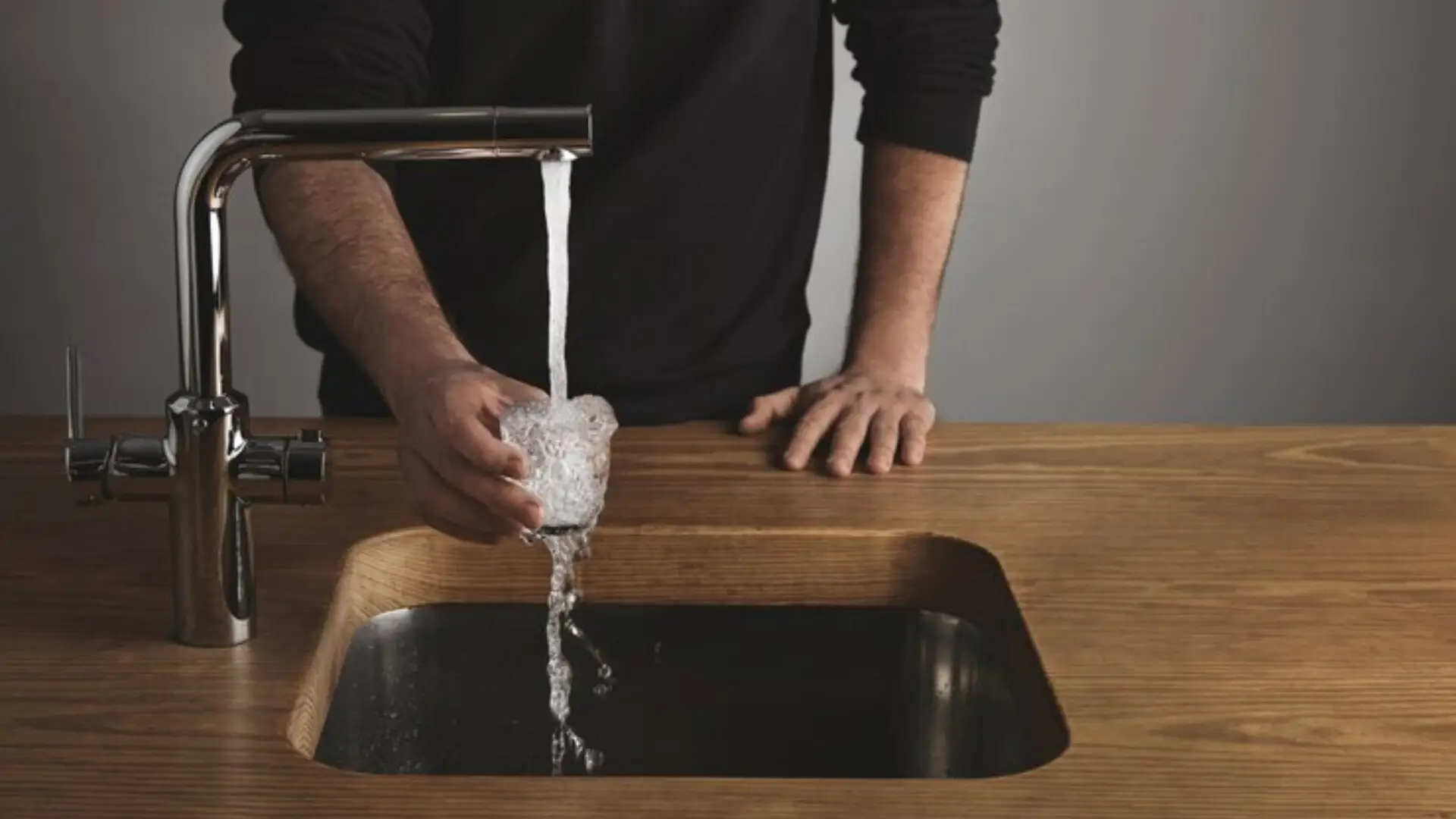 A person is holding a glass under a running faucet, letting water overflow into a modern wooden sink, mindful of Ofwat regulations.