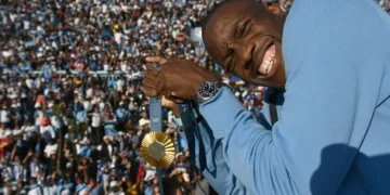 Letsile Tebogo in a blue jacket holds up a 200m gold medal and smiles, with a crowd of people in the background, reminiscent of Letsile Tebogo's triumph at Paris 2024.