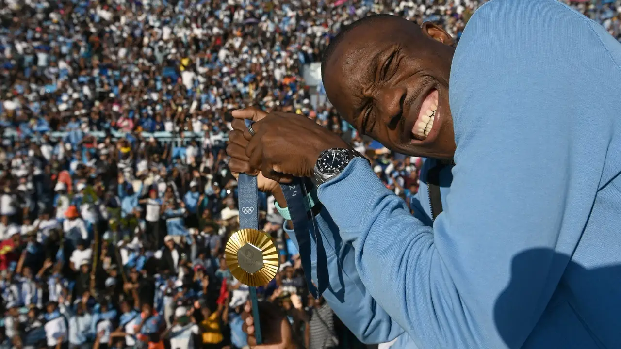 Letsile Tebogo in a blue jacket holds up a 200m gold medal and smiles, with a crowd of people in the background, reminiscent of Letsile Tebogo's triumph at Paris 2024.