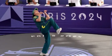 Rachael 'Raygun- Gunn in a green uniform performs a kick during a taekwondo event at the Paris 2024 Olympics, with judges seated at a table in the background.