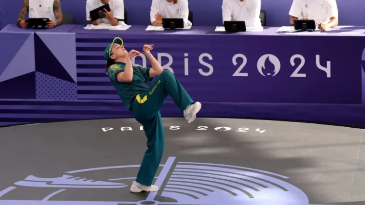 Rachael 'Raygun- Gunn in a green uniform performs a kick during a taekwondo event at the Paris 2024 Olympics, with judges seated at a table in the background.
