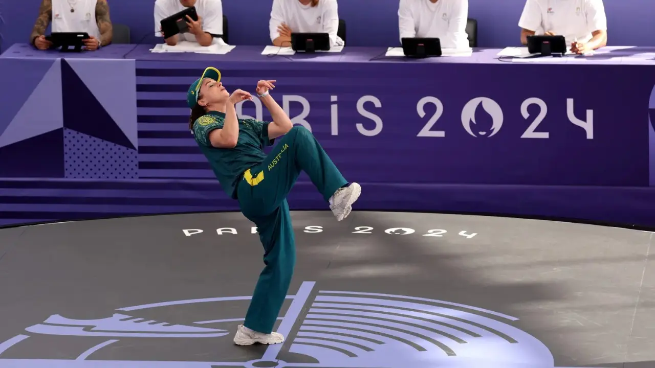 Rachael 'Raygun- Gunn in a green uniform performs a kick during a taekwondo event at the Paris 2024 Olympics, with judges seated at a table in the background.