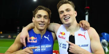Duplantis and Warholm in Puma sportswear, known for competing in the 100m Exhibition Sprint, stand side by side, smiling and pointing at each other's chests. One athlete wears a blue outfit, the other wears white. A night sky and sports field are in the background.