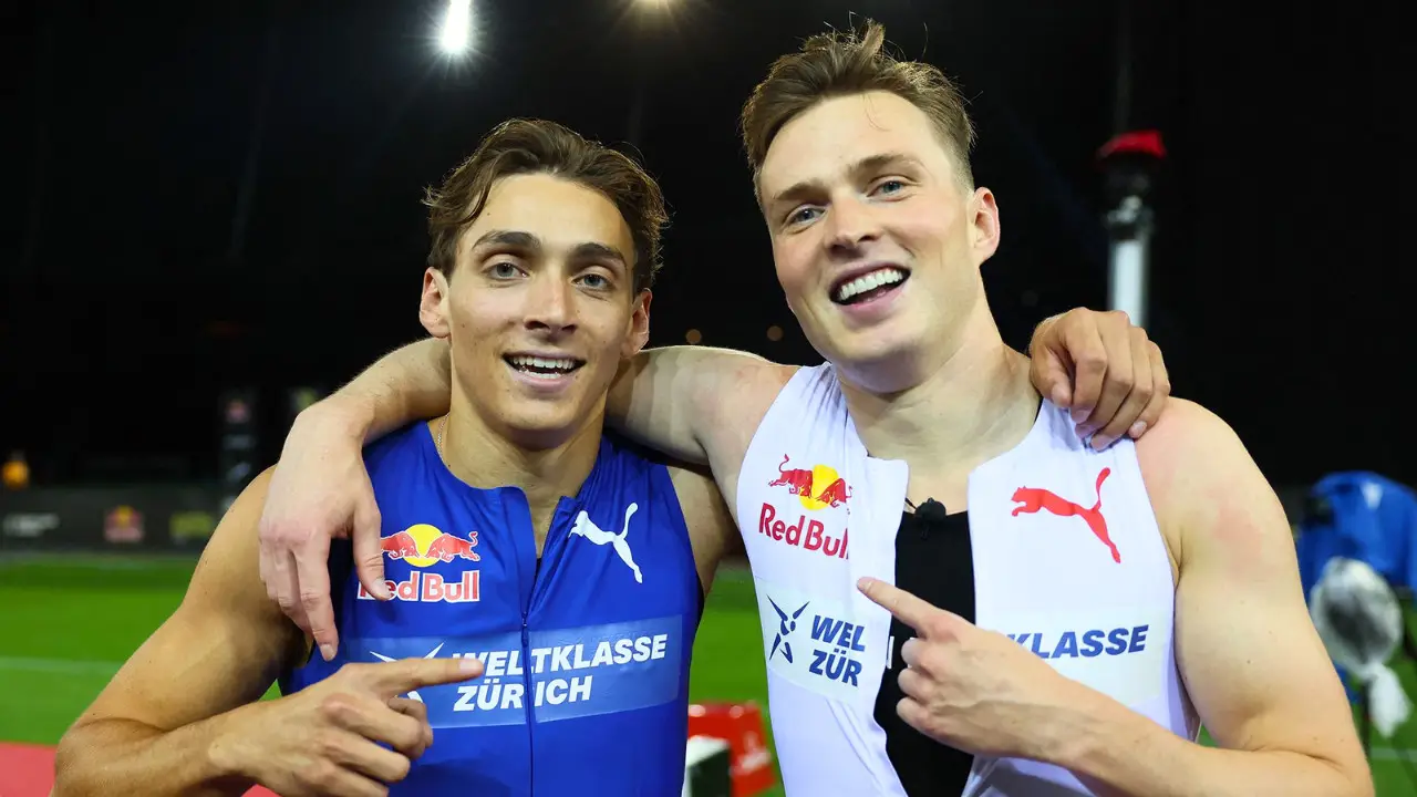 Duplantis and Warholm in Puma sportswear, known for competing in the 100m Exhibition Sprint, stand side by side, smiling and pointing at each other's chests. One athlete wears a blue outfit, the other wears white. A night sky and sports field are in the background.