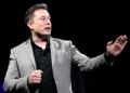 Elon Musk in a gray blazer and black shirt gestures while speaking against a dark background, envisioning the ambitious future where Elon Musk becomes the first trillionaire by 2027.