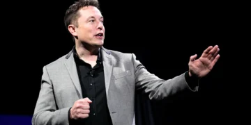 Elon Musk in a gray blazer and black shirt gestures while speaking against a dark background, envisioning the ambitious future where Elon Musk becomes the first trillionaire by 2027.