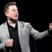 Elon Musk in a gray blazer and black shirt gestures while speaking against a dark background, envisioning the ambitious future where Elon Musk becomes the first trillionaire by 2027.