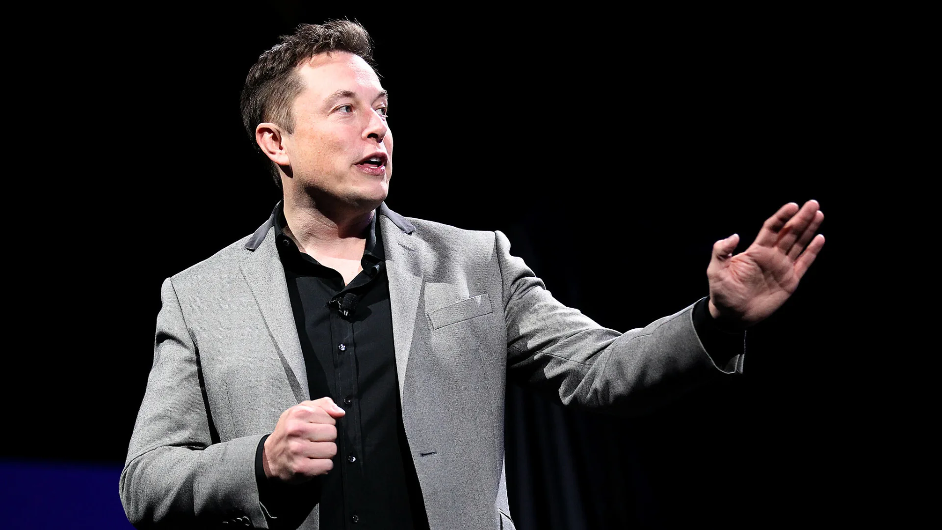 Elon Musk in a gray blazer and black shirt gestures while speaking against a dark background, envisioning the ambitious future where Elon Musk becomes the first trillionaire by 2027.