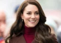A woman with long, dark brown hair smiles while wearing a maroon turtleneck and a brown coat in an outdoor setting, emanating the same grace and poise as Catherine, Princess of Wales.