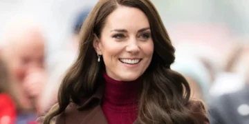 A woman with long, dark brown hair smiles while wearing a maroon turtleneck and a brown coat in an outdoor setting, emanating the same grace and poise as Catherine, Princess of Wales.