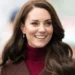 A woman with long, dark brown hair smiles while wearing a maroon turtleneck and a brown coat in an outdoor setting, emanating the same grace and poise as Catherine, Princess of Wales.
