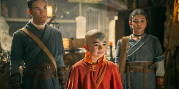 Three people dressed in detailed, fantasy-themed costumes stand side by side in a dimly lit room. The person in the middle has a distinct arrow tattoo on their bald head, reminiscent of Avatar: The Last Airbender.