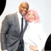 A man in a grey suit and a woman with pink hair in a white outfit stand closely together, both smiling at the camera. The background has circular inserts showing light designs of people. You might even mistake them for Terry Crews and his wife Rebecca sharing a joyful moment.