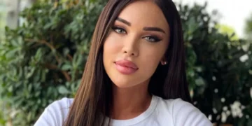 A woman with long brown hair and a white shirt stands in front of a leafy background. She has a neutral expression on her face, reflecting the ongoing struggles concerning Georgia LGBTQ rights.
