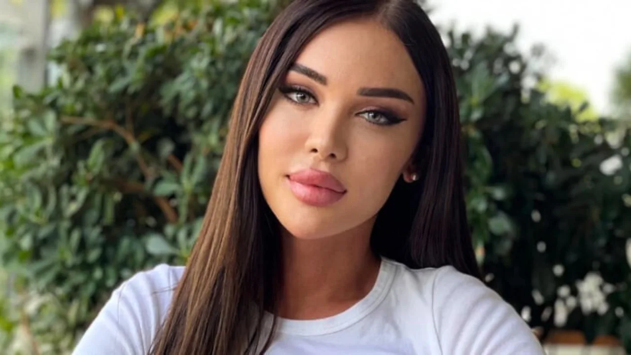 A woman with long brown hair and a white shirt stands in front of a leafy background. She has a neutral expression on her face, reflecting the ongoing struggles concerning Georgia LGBTQ rights.