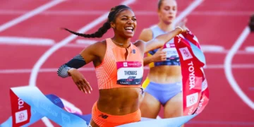 Gabby in orange uniform crosses the finish line with a smiling expression, holding a finish tape at the ATHLOS NYC inaugural track event. Another athlete in the background.