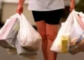 A person wearing a white shirt and black shorts is seen carrying multiple grocery bags in both hands, mindful of California's plastic bag ban aimed at plastic waste reduction.