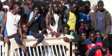 A large group of people stand closely together behind a white barrier, some looking towards the camera while others look away, perhaps pondering EU asylum policies. They are outdoors on what appears to be a sunny day.