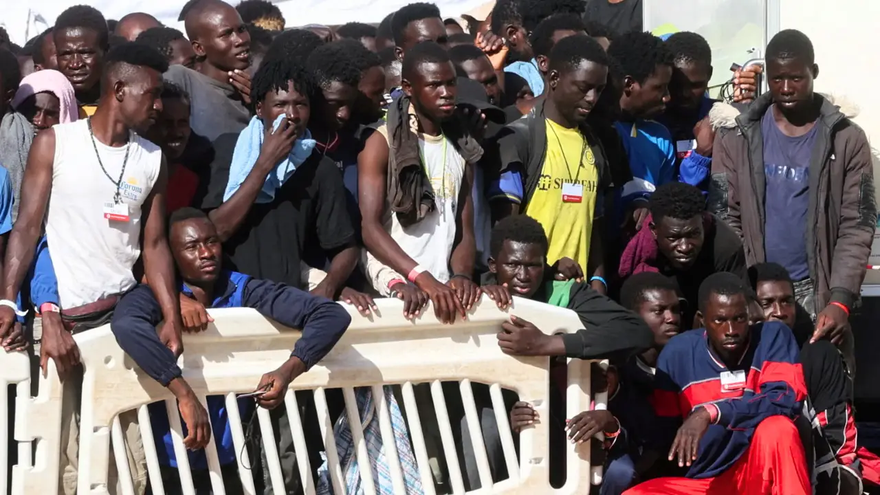 A large group of people stand closely together behind a white barrier, some looking towards the camera while others look away, perhaps pondering EU asylum policies. They are outdoors on what appears to be a sunny day.