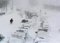 A snowy street with numerous cars buried under snow, some people walking around, and heavy snowfall continuing steadily.
